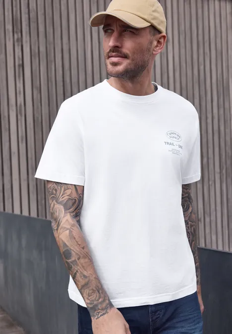 Een man in een wit T-shirt met opvallende armtattoos staat voor een houten lattenwand.