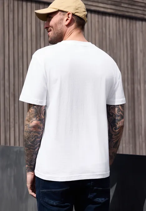 Man van achteren met beige pet, wit T-shirt, donkere jeans en getatoeëerde armen voor een houten wand.