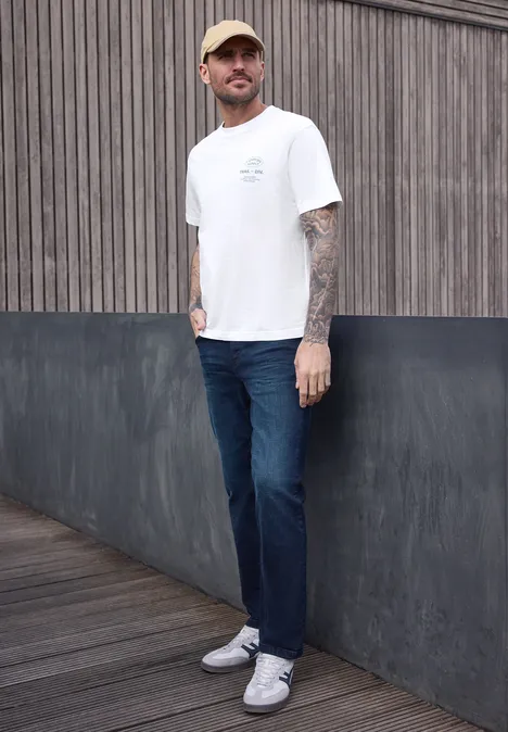 Een getatoeëerde man draagt een wit t-shirt, blauwe jeans en witte sneakers voor een houten en metalen wand.