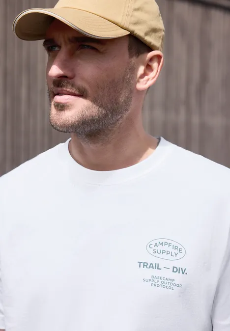 Man met pet en wit T-shirt met logo voor een houten wand.