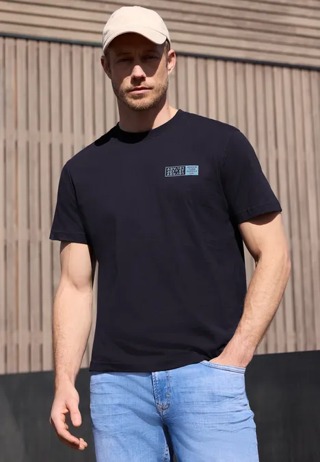 Een man in zwart T-shirt en blauwe jeans staat voor een houten wand.