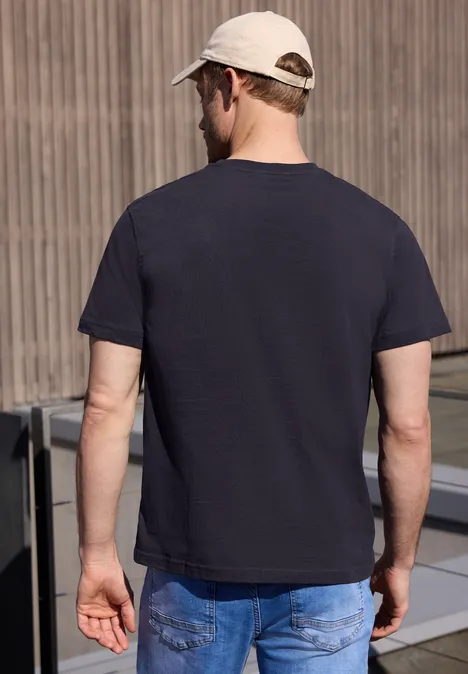 Een man van achteren gezien draagt een donkerblauw t-shirt, blauwe spijkerbroek en een lichte pet voor een muur.