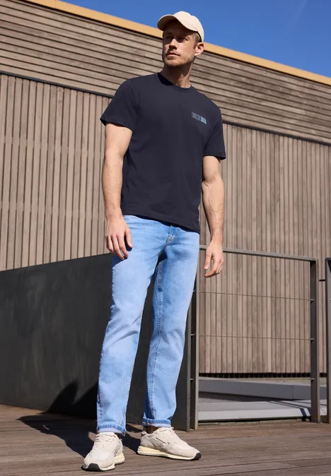 Een man in een zwart T-shirt, lichtblauwe spijkerbroek en witte sneakers staat buiten voor een houten gevel.