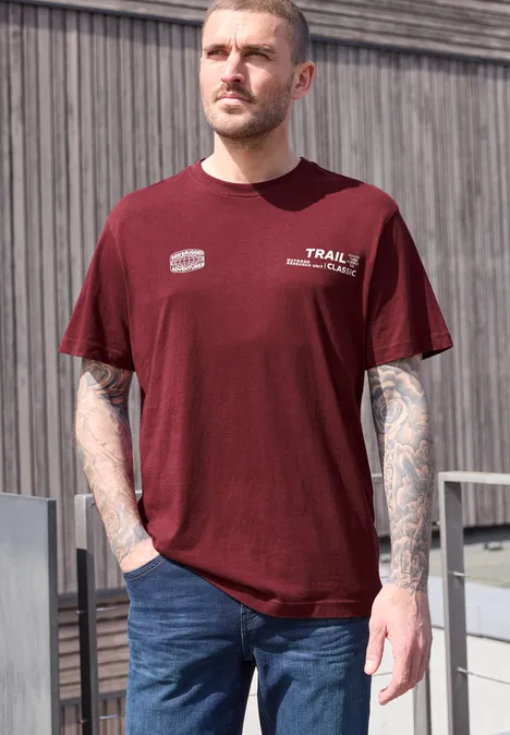 Man staat buiten, draagt bordeauxrood t-shirt, spijkerbroek en heeft getatoeëerde armen.