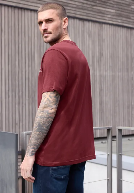 Jonge man met tatoeages in rood T-shirt staand zijwaarts voor een houten gevel.