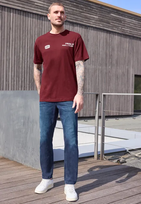 Een man met een rood t-shirt, blauwe jeans en witte sneakers staat op een houten terras voor een gebouw.