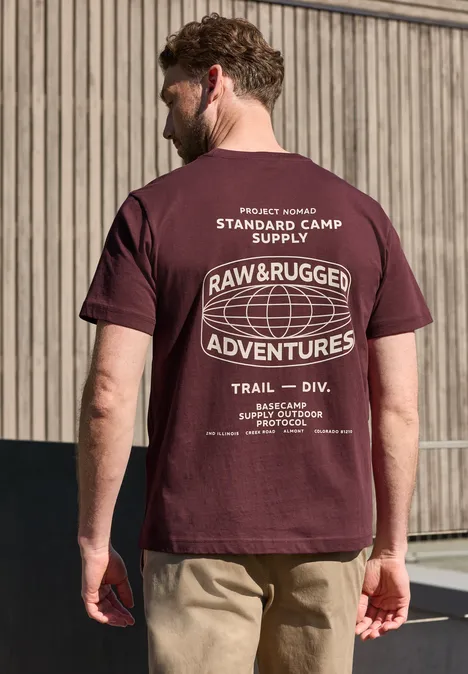Een man in een beige broek voor een houten gevel draagt van achteren een bordeaux T-shirt met 'Raw & Rugged Adventures'-opdruk.