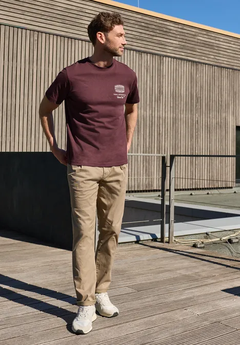 Een man staat op een houten terras, draagt een bordeaux T-shirt, beige broek en witte sneakers.