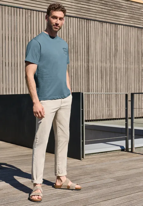Een man staat ontspannen op een houten terras voor een lattenwand, draagt een blauw T-shirt, lichte broek en sandalen.