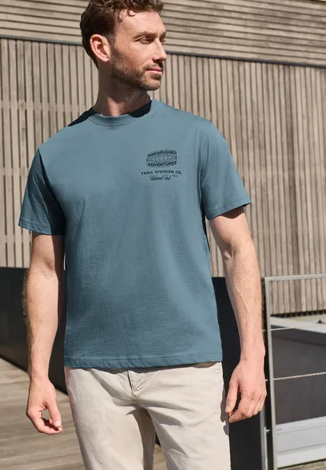 Een man in een blauwgroen T-shirt en lichte broek voor een moderne houten gevel.