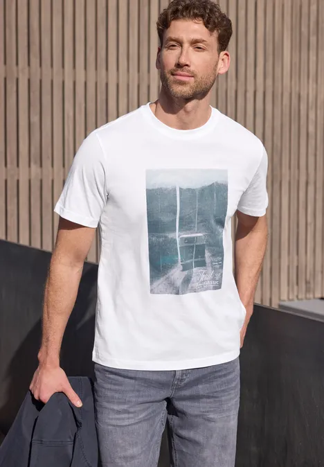Man voor houten wand, draagt wit bedrukt t-shirt, blauwe jeans en houdt een donkere jas vast.