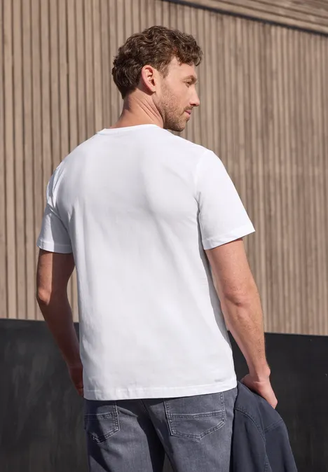 Een man staat met een wit t-shirt en spijkerbroek voor een houten wand en houdt een jas in zijn hand.