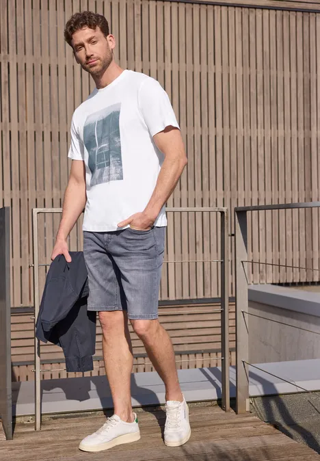 Een man in een wit t-shirt en grijze short staat op een balkon met een jas in de hand.