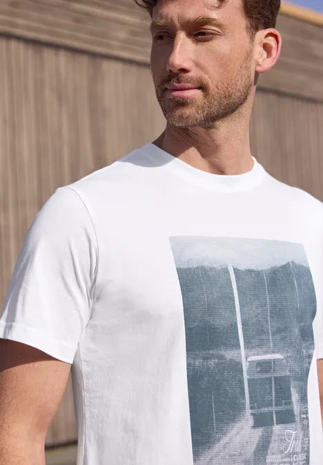 Een bebaarde man draagt een wit T-shirt met berglandschap en kabelbaan voor een houten wand.