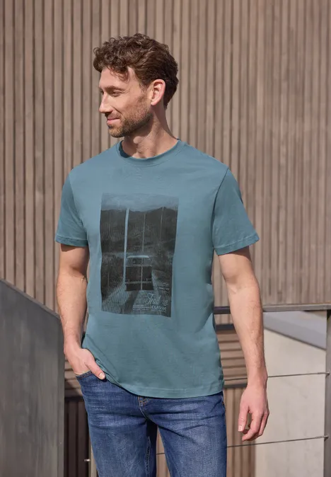 Een man in een turquoise T-shirt met print en spijkerbroek staat nonchalant voor een houten muur.