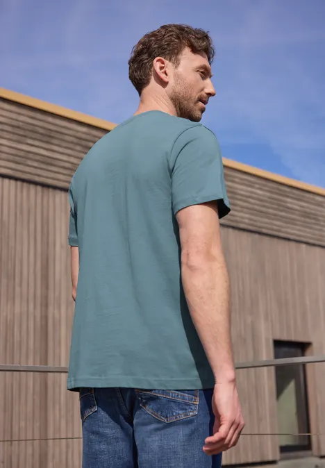 Een man met zijn rug naar de camera draagt een turquoise t-shirt en spijkerbroek voor een houten gevel.