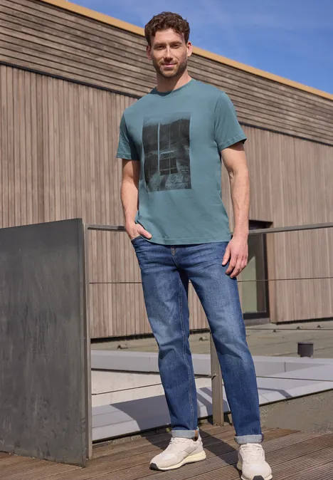 Een man in een groen T-shirt met print, blauwe jeans en sneakers staat op een betonnen terras voor een houten gevel.