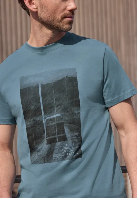 Zijdelings afgebeelde man in een turquoise t-shirt met zwarte rechthoekige print op de borst.