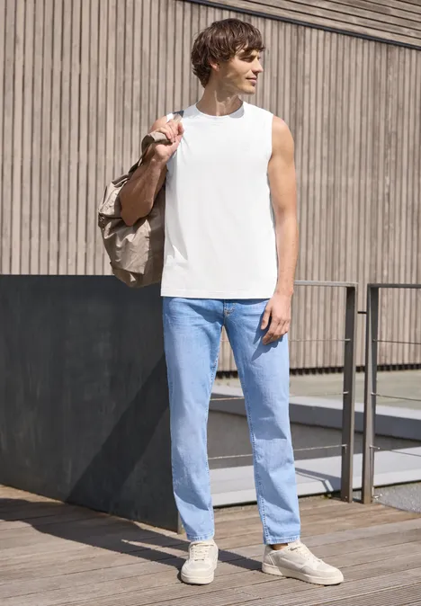 Een man staat voor een houten gevel, draagt een wit mouwloos shirt, lichte spijkerbroek, beige sneakers en een tas.