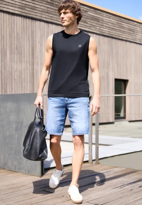 Een man in een zwart tanktop, spijkershorts en witte sneakers, met een zwarte tas op een houten terras.