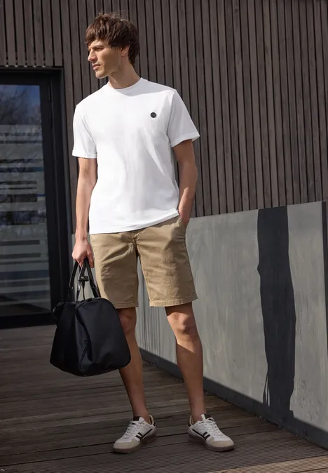 Een man in wit T-shirt, beige short en witte sneakers houdt een zwarte tas vast voor een moderne gevel.