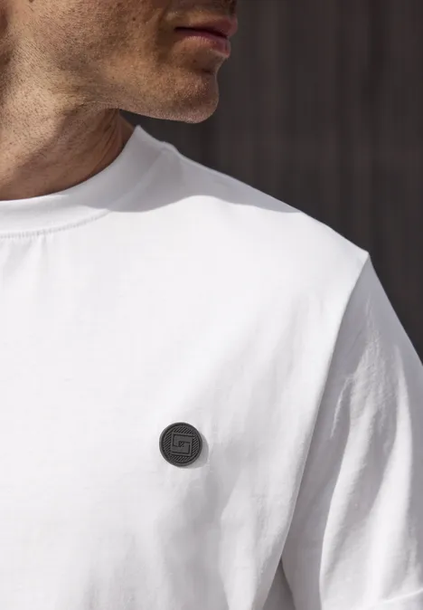 Een man in een wit t-shirt met een rond zwart embleem op de borst.