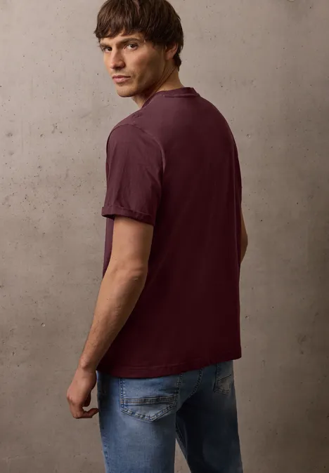 Een man staande opzij voor een grijze muur, gekleed in een donkerrode polo en blauwe spijkerbroek.