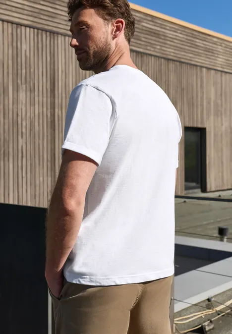 Een man met zijn rug naar de camera op een terras, draagt een wit t-shirt en kaki broek voor een houten gevel.