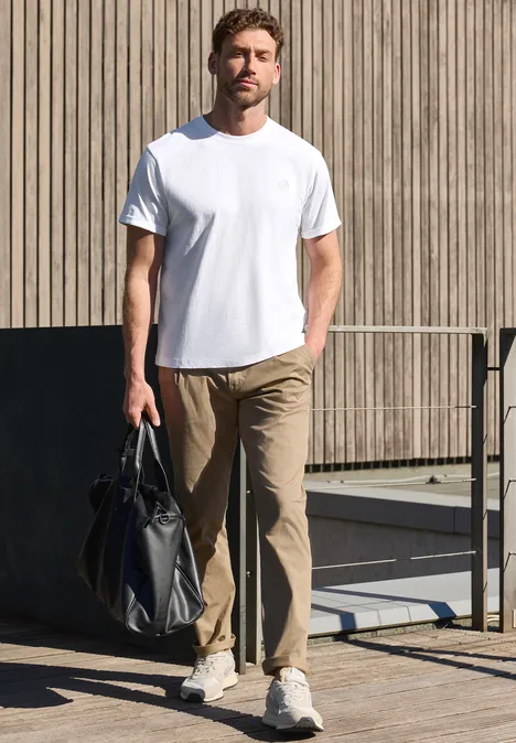 Man in wit T-shirt, beige broek en sneakers met een tas voor een houten wand.