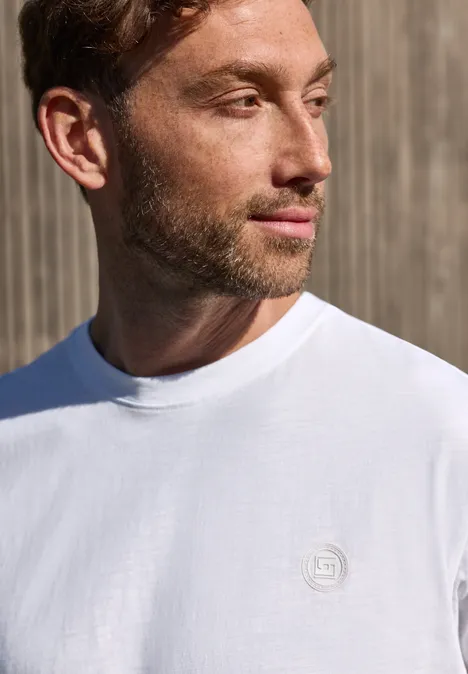 Een man in een wit T-shirt met klein logo voor een houten muur
