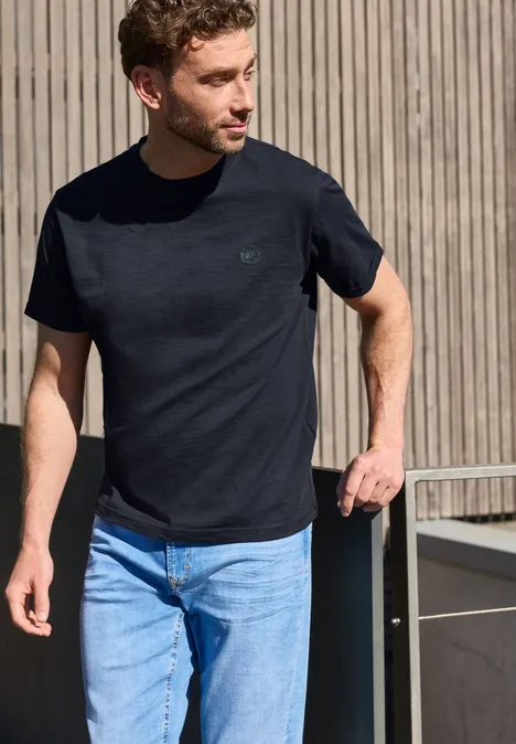 Een man in een zwart T-shirt en blauwe jeans staat nonchalant voor een houten wand.