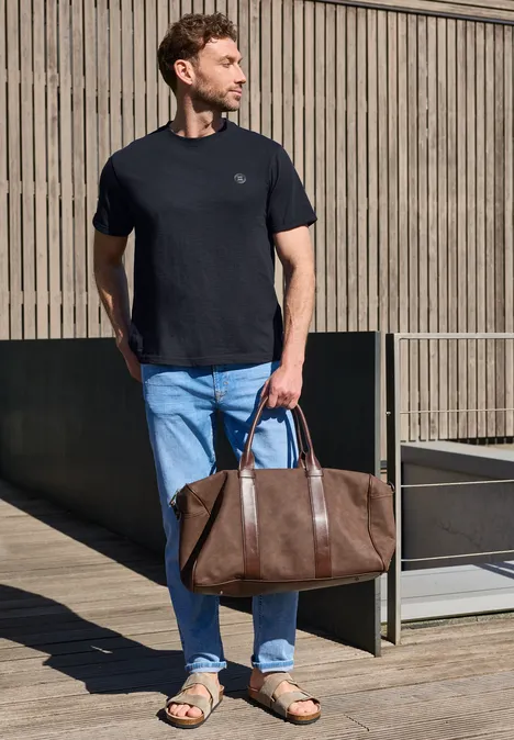 Een man in een zwart T-shirt en spijkerbroek houdt een bruine leren reistas vast voor een houten muur.