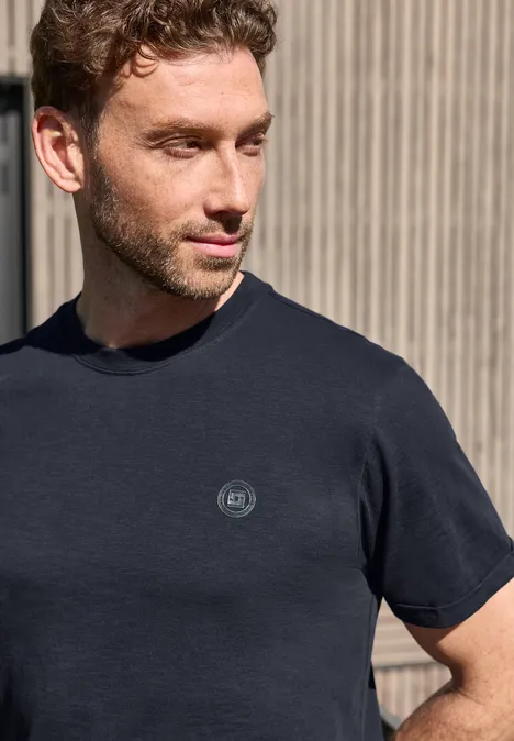 Een man in een donker T-shirt met klein logo staat voor een muur met verticale latten.