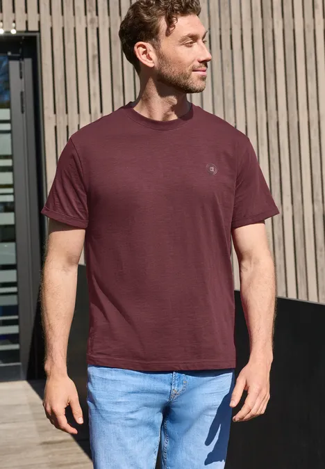 Een man in een wijnrood T-shirt en spijkerbroek staat buiten voor houten panelen.