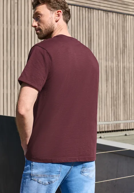 Een man in bordeauxrood T-shirt en blauwe spijkerbroek, van opzij en achterkant gefotografeerd voor een houten wand.