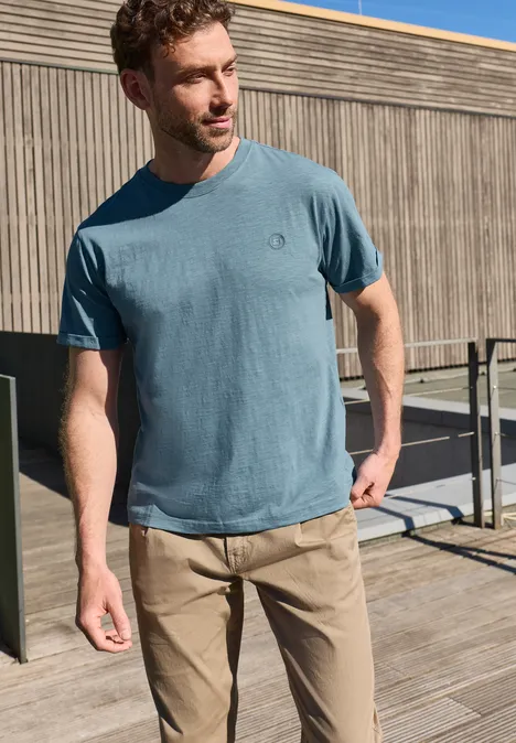 Een man in een blauw T-shirt en beige broek staat op een houten terras voor een houten gevel.