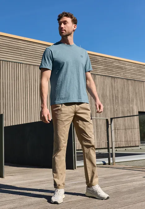 Man in blauw T-shirt en beige broek staat voor een moderne houten gevel.