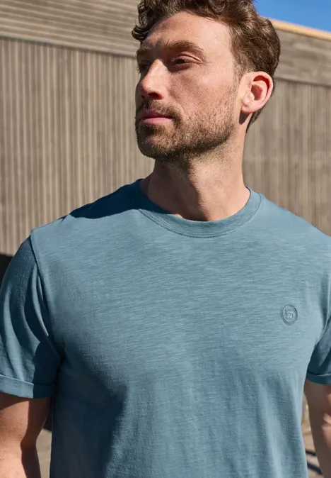 Man staat voor een houten wand in een lichtblauw T-shirt en kijkt opzij.