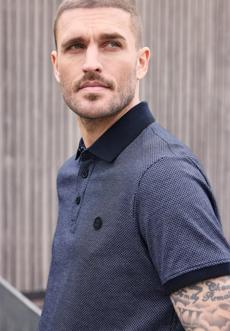 Ein Mann in blauem Poloshirt steht vor einer Holzwand, sichtbarer Unterarm mit Tattoo.