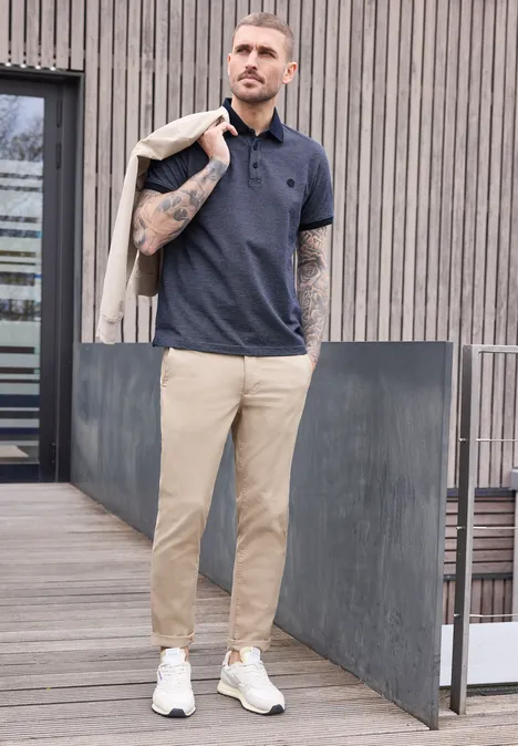 Ein tätowierter Mann in Poloshirt, beigen Chinos und weißen Schuhen trägt eine Jacke über der Schulter.