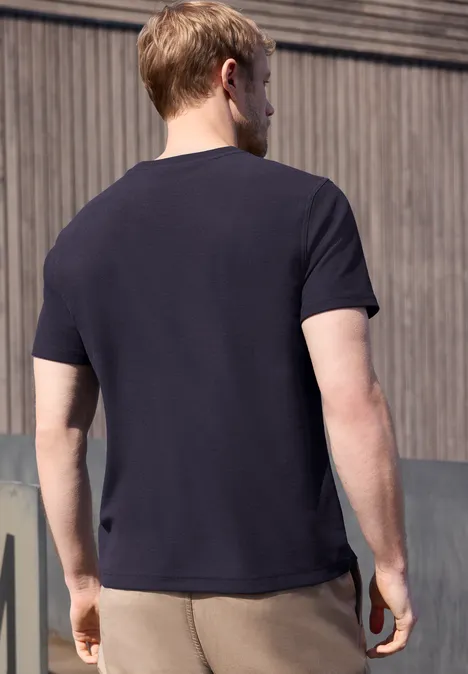 Ein Mann steht draußen, Rücken zur Kamera, in dunkelblauem T-Shirt und beigefarbener Hose vor Holzwand.