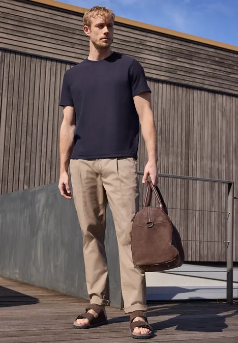 Ein Mann steht vor einer Holzfassade in schwarzem T-Shirt, beigen Hosen und Sandalen, hält eine braune Tasche.