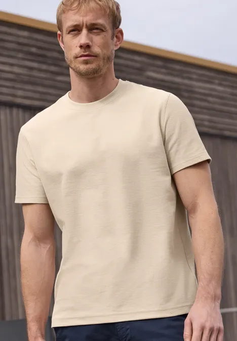 Ein Mann trägt ein schlichtes beiges T-Shirt vor einem Holzhintergrund.