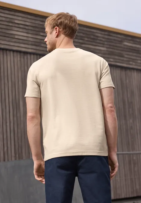 Ein Mann steht mit dem Rücken zur Kamera und trägt ein beiges T-Shirt sowie dunkle Hosen vor einer Holzfassade.