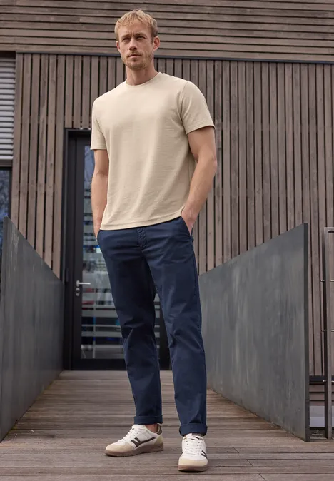 Ein Mann steht vor einer modernen Holzwand und trägt ein beiges T-Shirt, blaue Hose und weiße Sneaker.