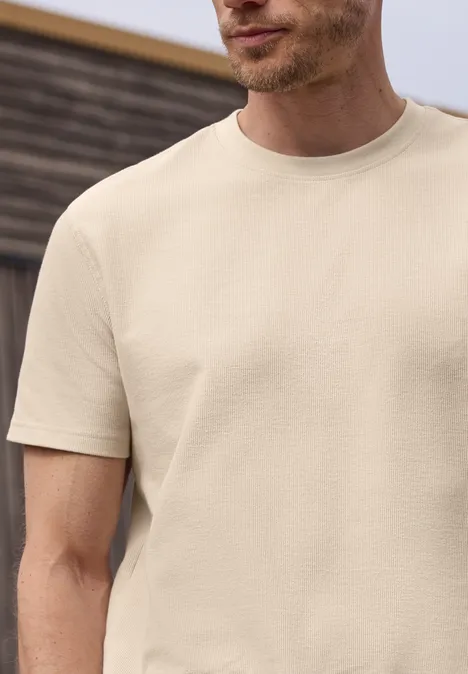 Ein Mann trägt ein beiges Kurzarm-T-Shirt vor unscharfem Holzhintergrund.