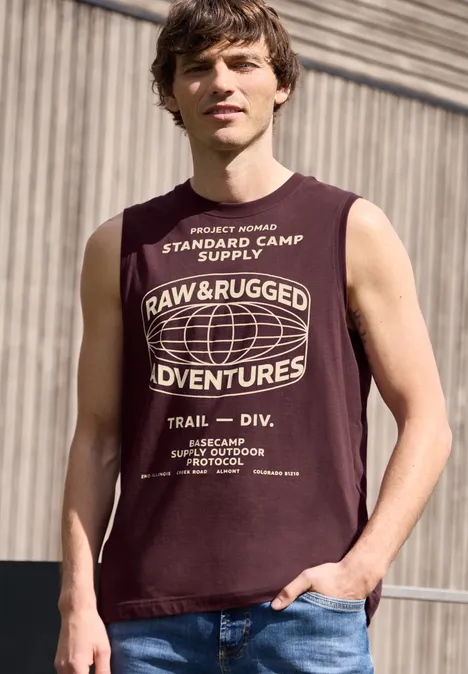 Een man draagt een donkerrood mouwloos T-shirt met witte opdruk 'Raw & Rugged Adventures' op industriële achtergrond.