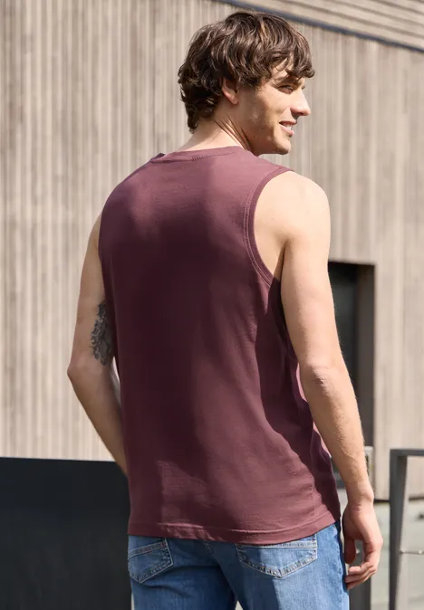 Een man van achteren gefotografeerd in een bordeaux tanktop en spijkerbroek voor een houten gevel.