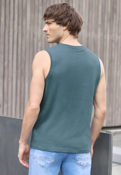 Een man staat met zijn rug naar de camera en draagt een petrolkleurige tanktop voor een houten muur.