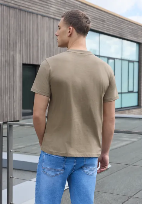 Shirt met ronde hals en Wording Artwork antique beige Een man in beige T-shirt en spijkerbroek staat op een dakterras en kijkt naar een moderne glazen gevel.